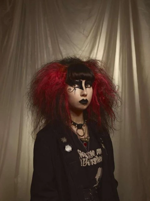 Portrait of a woman with goth makeup and vibrant red and black hair, wearing a black jacket, against a textured beige backdrop.