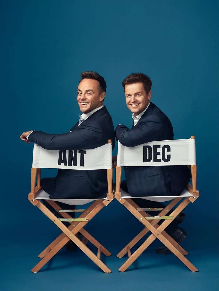 Ant & Dec seated back-to-back on director's chairs in a studio, both wearing dark suits and smiling, set against a blue backdrop.