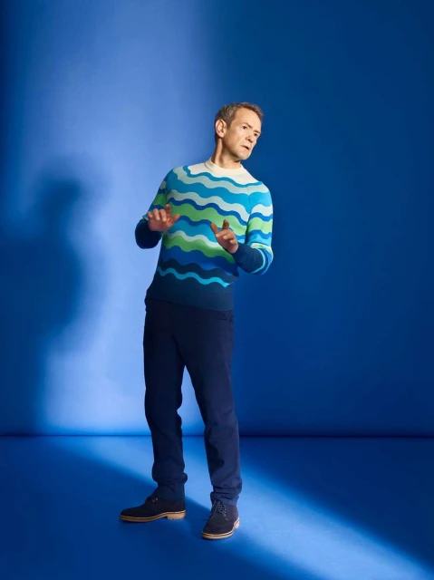 Adult male in a blue studio, wearing a patterned sweater and dark pants, looking to the side and gesturing with his hands.