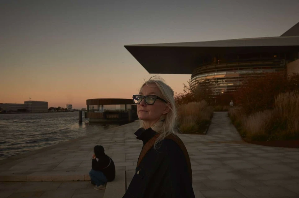Woman in glasses by waterfront architecture at dusk. Modern building reflects sunset. Another person sits on the ledge.