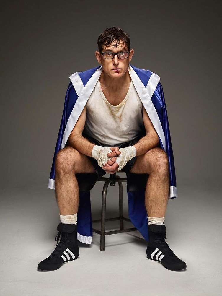 A male boxer in a blue and white robe sits on a stool with his hands wrapped, looking at the camera in an indoor setting.