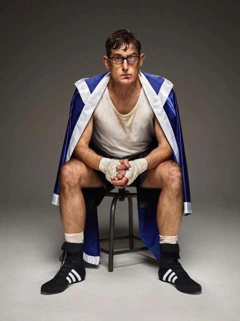 A male boxer in a blue and white robe sits on a stool with his hands wrapped, looking at the camera in an indoor setting.