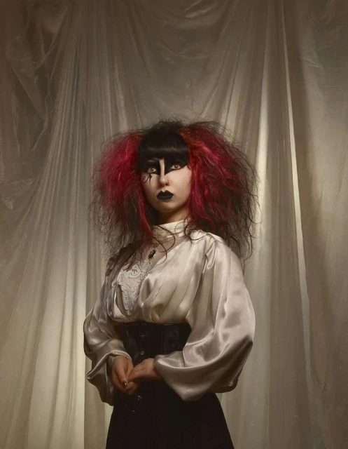 A young woman with vivid makeup and distinctive red-tipped hairstyle strikes a pose indoors against a textured white backdrop. She is wearing a satin blouse and corset.