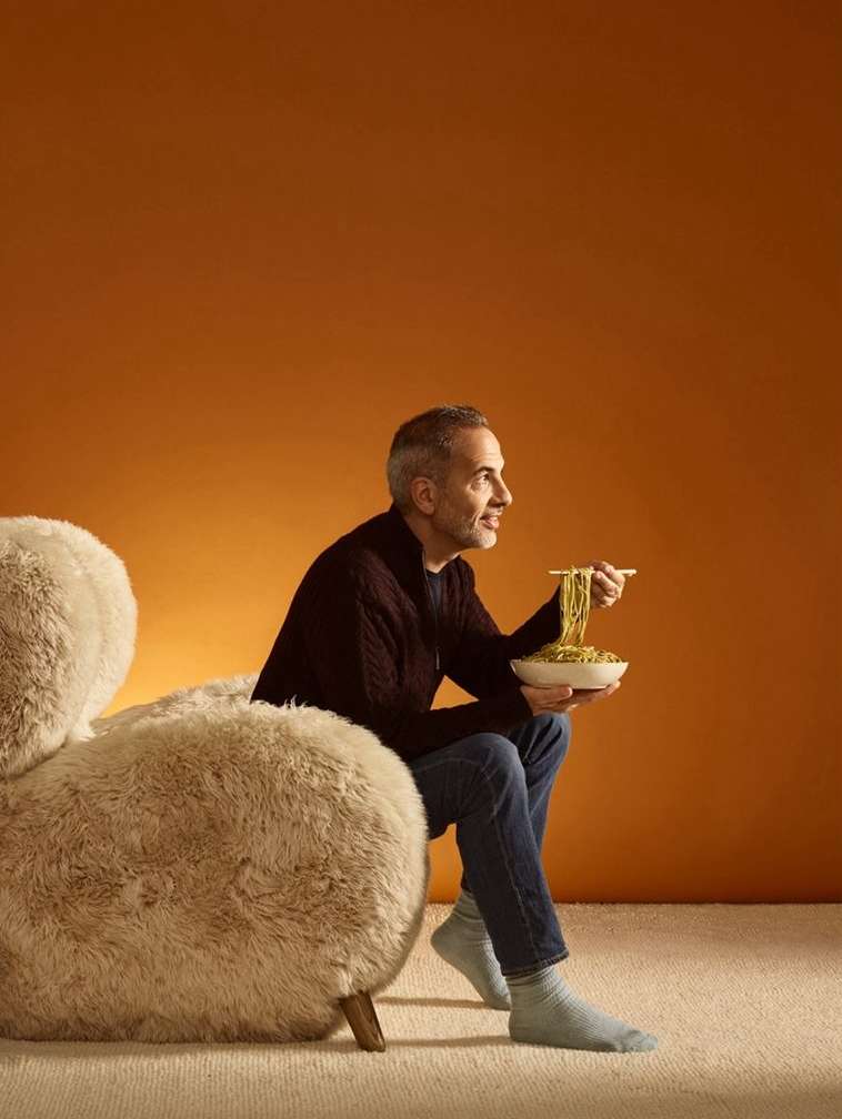 Mature man with gray hair enjoys noodles with chopsticks while seated on a fuzzy chair against an orange backdrop in a warm indoor setting.