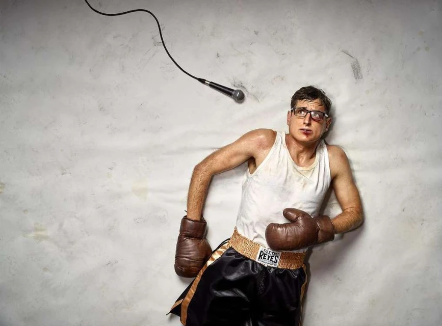 Man in boxing attire lies on ground next to microphone. He wears boxing gloves, black shorts with 'REYES' on the waistband.