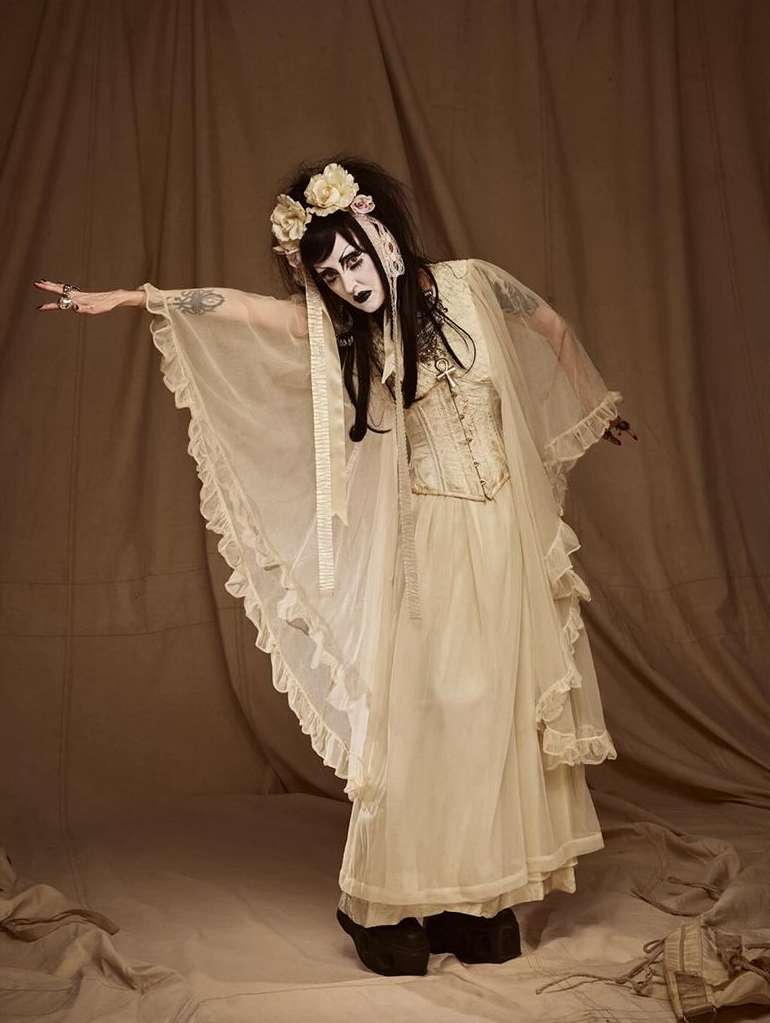 A person with theatrical makeup and an elaborate floral headband poses in a flowing gown with a vintage corset in a studio setting.