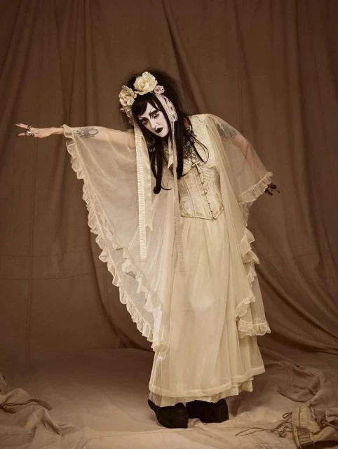 A person with theatrical makeup and an elaborate floral headband poses in a flowing gown with a vintage corset in a studio setting.