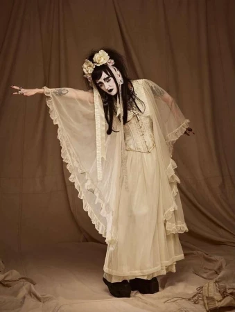 A person with theatrical makeup and an elaborate floral headband poses in a flowing gown with a vintage corset in a studio setting.