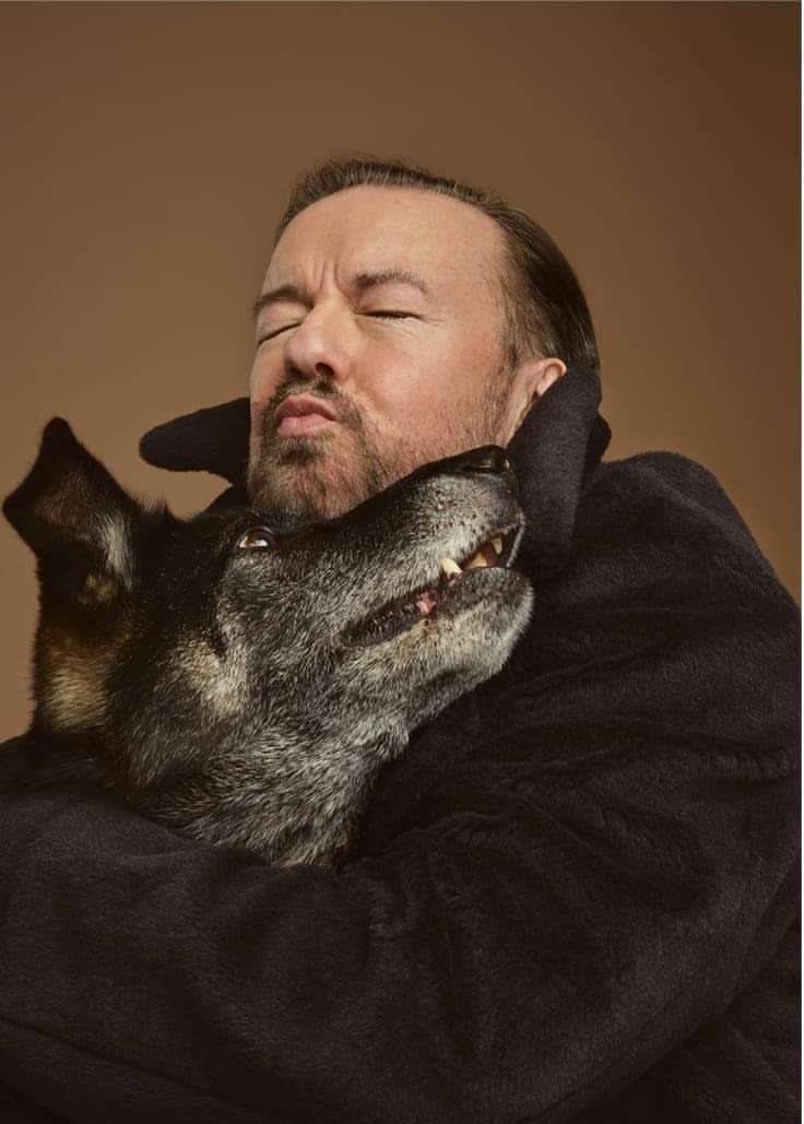 Portrait of Ricky Gervais with his eyes closed hugging his dog. The background is light brown. He is wearing a dark robe.