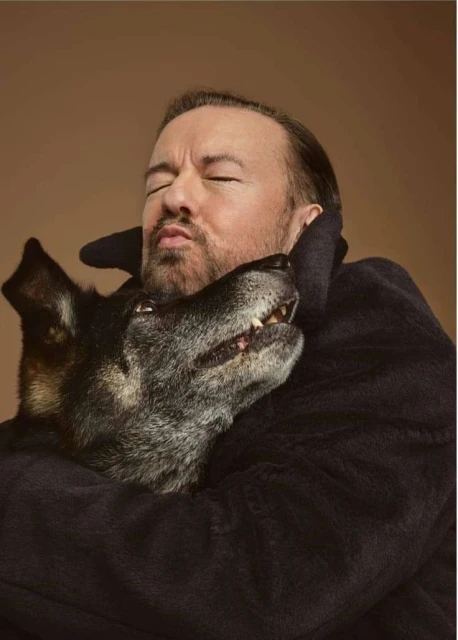 Portrait of Ricky Gervais with his eyes closed hugging his dog. The background is light brown. He is wearing a dark robe.