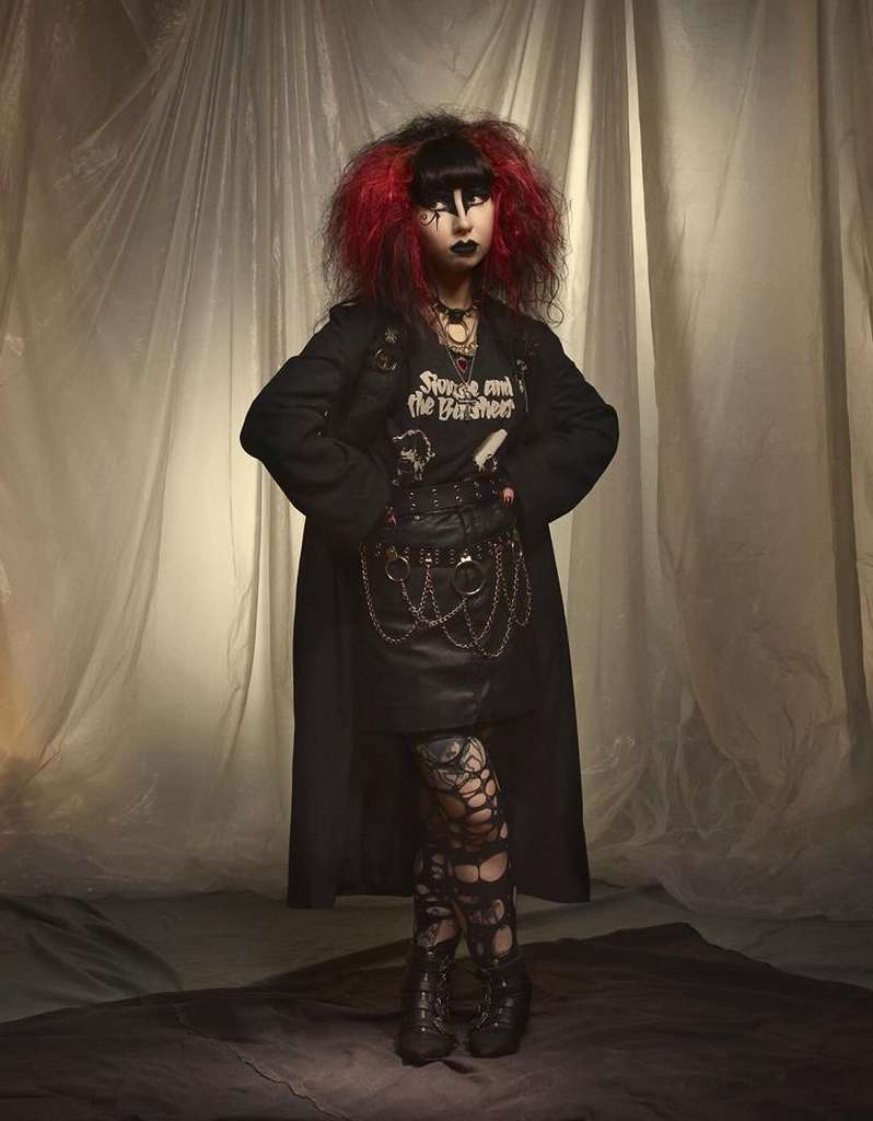 Full-length portrait of a woman in a goth-inspired outfit with dark makeup and dyed red-tipped hair in front of a draped background.