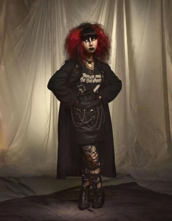 Full-length portrait of a woman in a goth-inspired outfit with dark makeup and dyed red-tipped hair in front of a draped background.