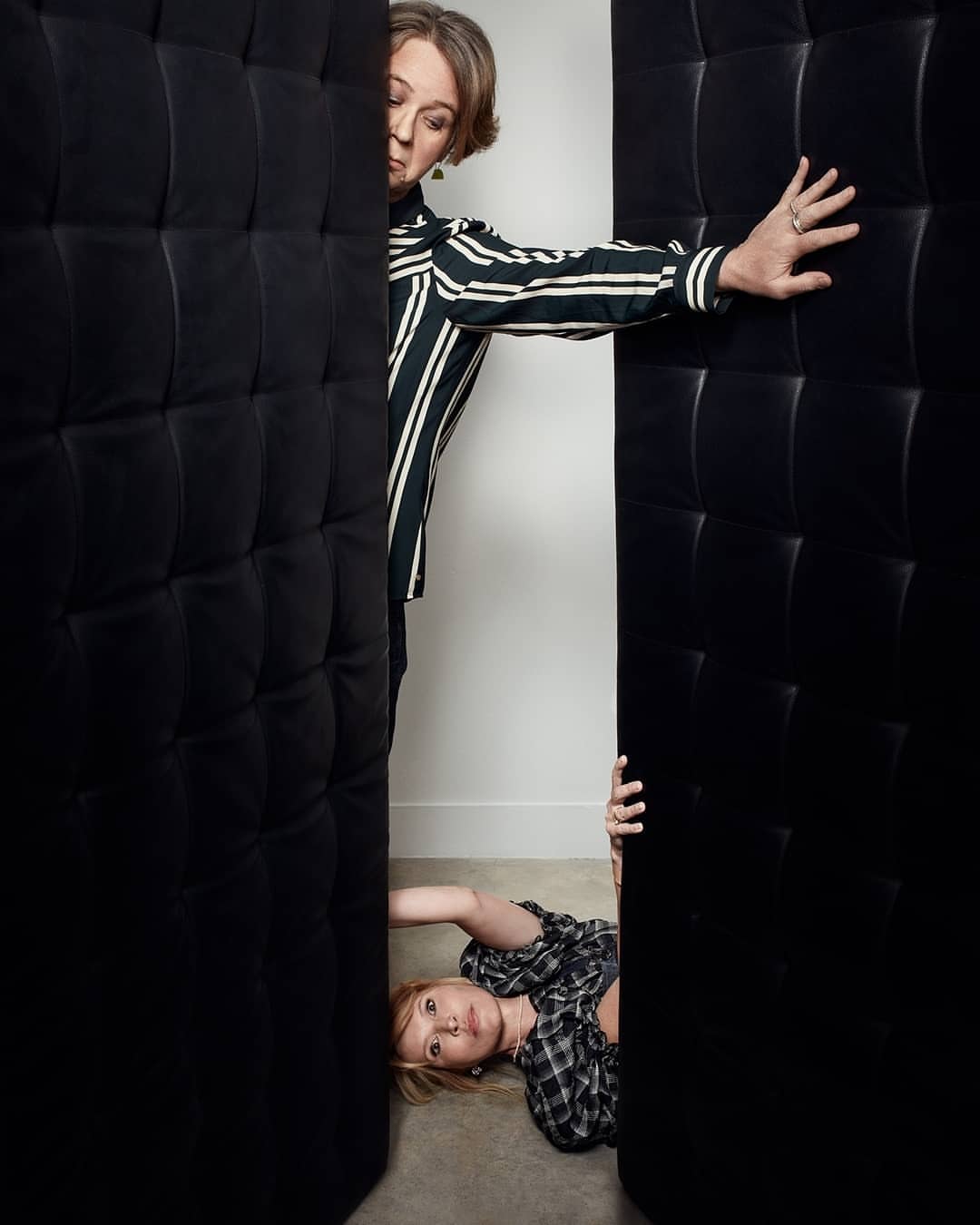 Two women pose between dark cushioned panels. One woman in stripes stands, while a second woman in plaid reclines on the floor.
