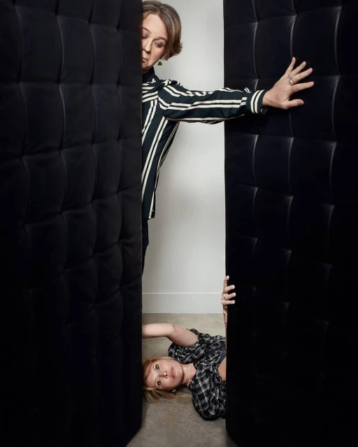 Two women pose between dark cushioned panels. One woman in stripes stands, while a second woman in plaid reclines on the floor.