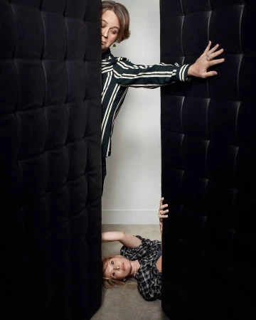 Two women pose between dark cushioned panels. One woman in stripes stands, while a second woman in plaid reclines on the floor.