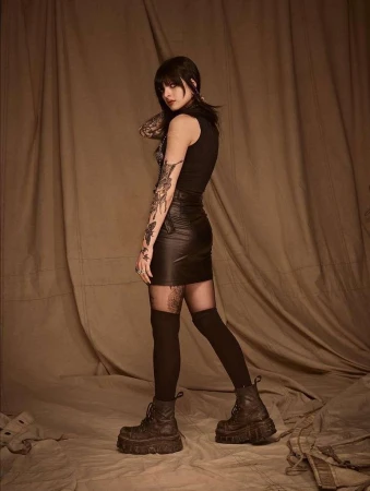 Woman in black sleeveless top and leather mini skirt standing against draped beige fabric backdrop