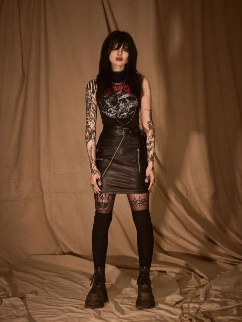 Full-length portrait of a young woman with dark hair and tattoos wearing a graphic top and leather skirt in a photo studio.