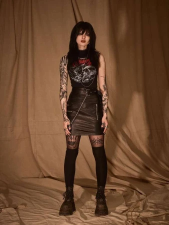 Full-length portrait of a young woman with dark hair and tattoos wearing a graphic top and leather skirt in a photo studio.
