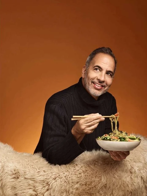 Smiling adult man holding chopsticks and bowl of salad with noodles on a furry surface against an orange studio backdrop.