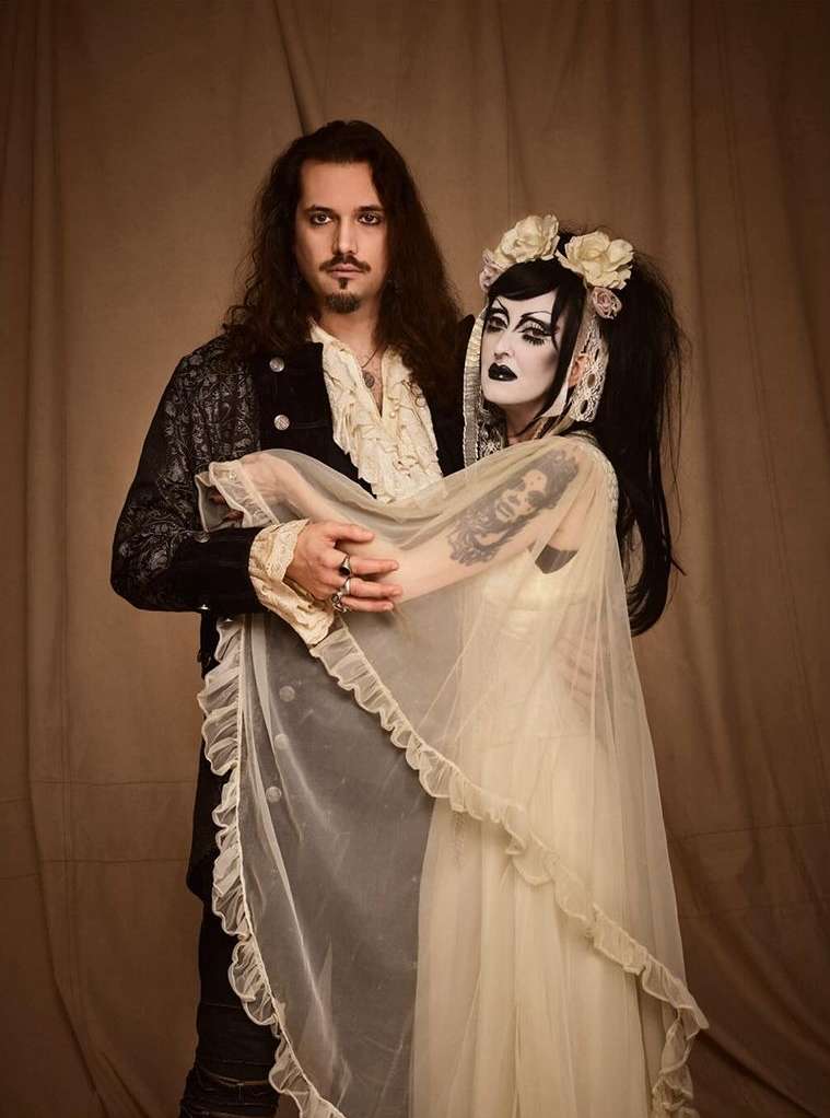 Gothic couple in formalwear strike a dramatic pose in a studio setting, the woman adorned with a veil and floral crown, against a tan backdrop.