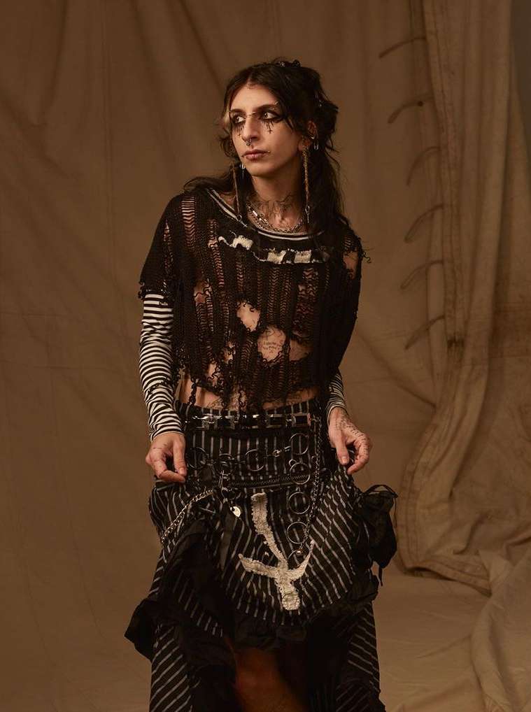 Young adult female fashion model posing in distressed black top, striped shirt and skirt, with dark eye makeup, in a studio setting.