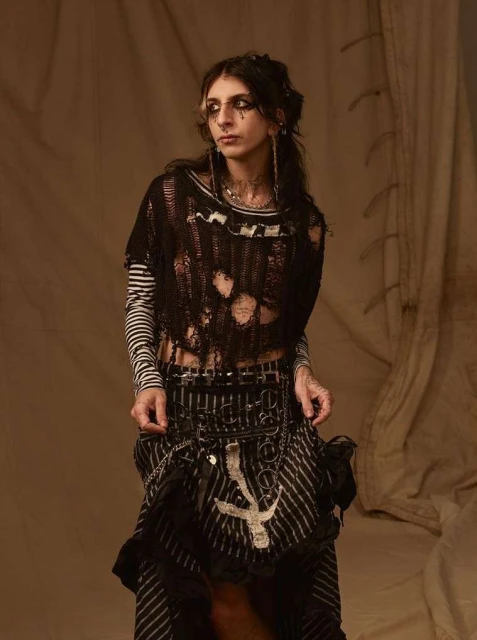 Young adult female fashion model posing in distressed black top, striped shirt and skirt, with dark eye makeup, in a studio setting.