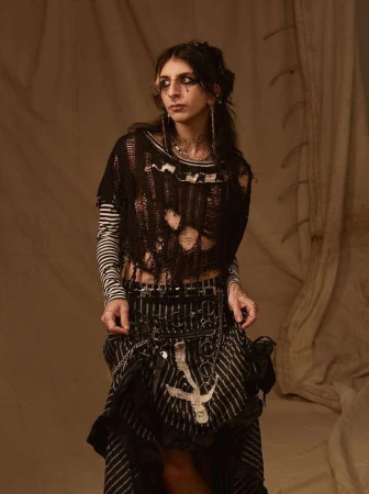Young adult female fashion model posing in distressed black top, striped shirt and skirt, with dark eye makeup, in a studio setting.