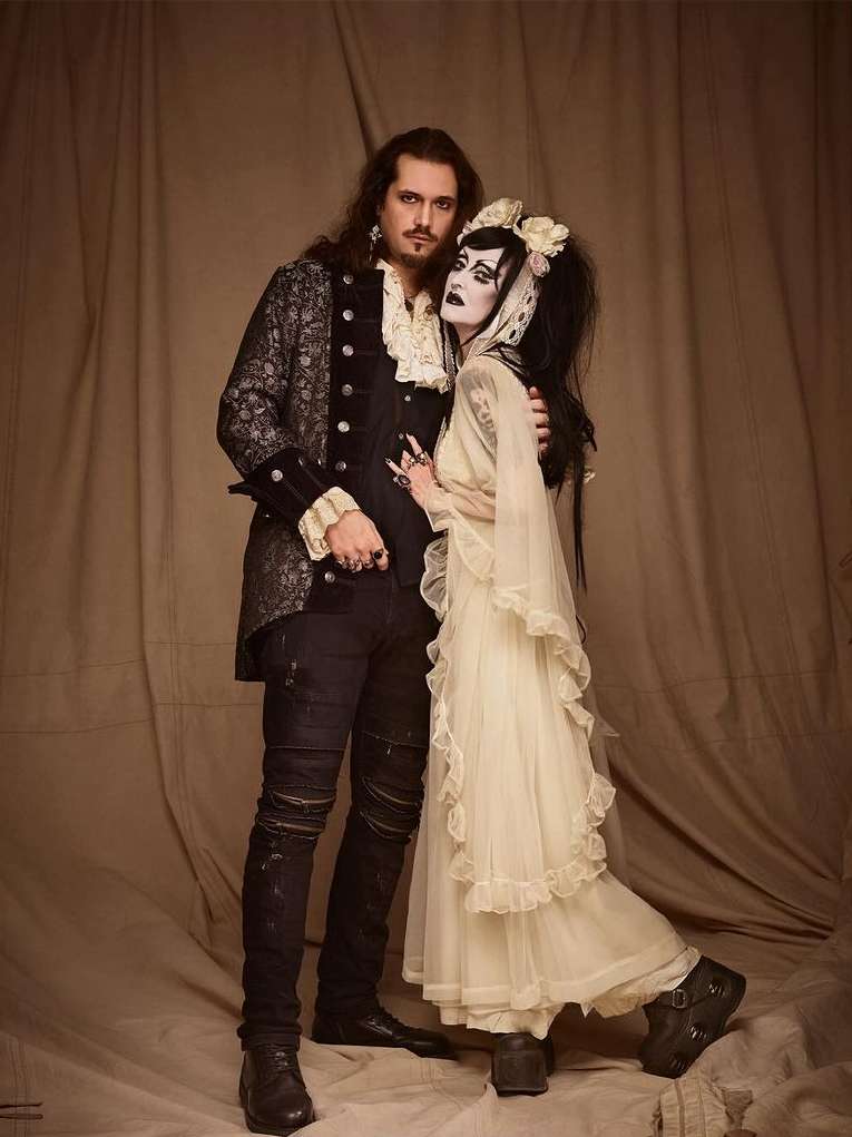 A vintage-style photo of a gothic couple embracing in a photo studio with dramatic makeup, period clothing, and a neutral backdrop.