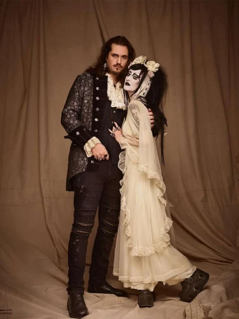A vintage-style photo of a gothic couple embracing in a photo studio with dramatic makeup, period clothing, and a neutral backdrop.
