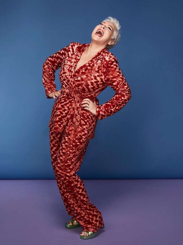Full shot of an adult woman laughing in a patterned red velvet suit on a blue and lavender background in a photo studio.