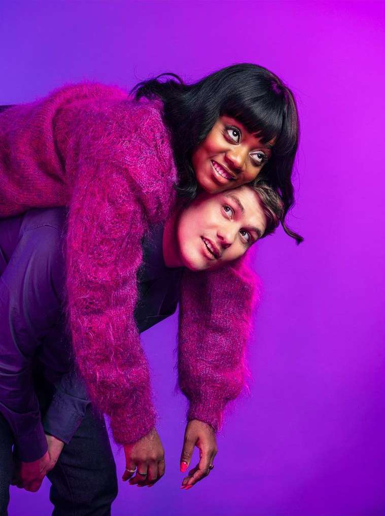 A smiling woman rides piggyback on a smiling man against a purple background in a medium shot, wearing casual outfits and jewelry.