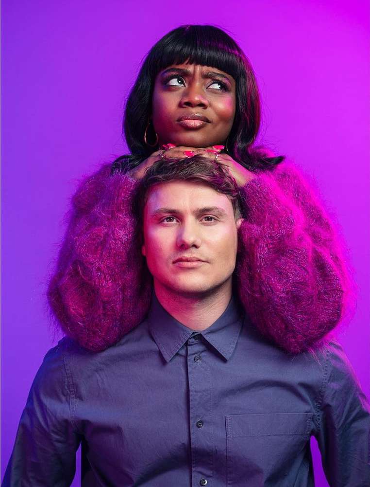 Portrait of a woman with a neutral expression holding a man's head, both indoors against a purple background with soft, diffused lighting.
