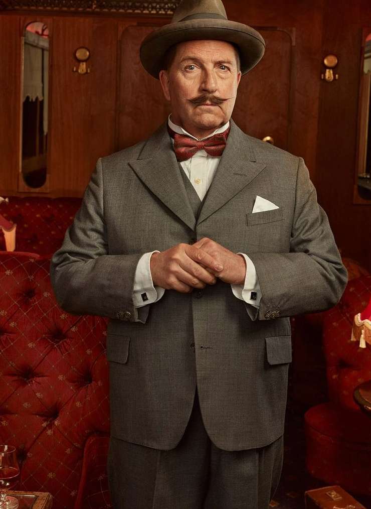 Portrait of a mature man in a grey suit, bow tie, and fedora, standing in an ornate, vintage train car with red velvet seating.