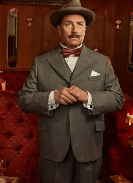 Portrait of a mature man in a grey suit, bow tie, and fedora, standing in an ornate, vintage train car with red velvet seating.