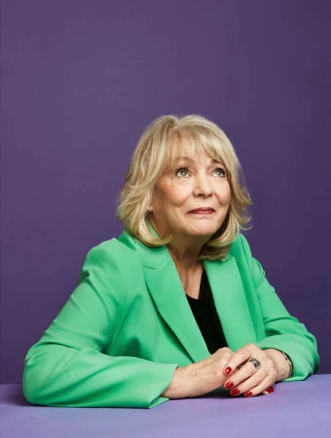 Mature woman in a green blazer poses against a purple background, looking upward with her hands clasped on a matching purple table.