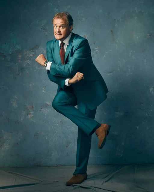 Man in a teal suit strikes a dynamic pose with a confident expression against a mottled grey and blue backdrop. 
