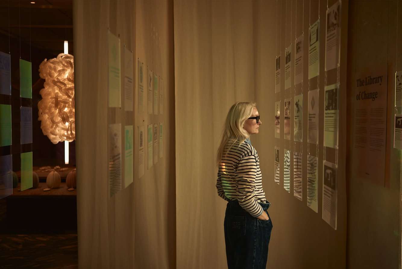 Woman in striped shirt examines informational panels at gallery exhibit with off-white walls and unique modern light fixture nearby.