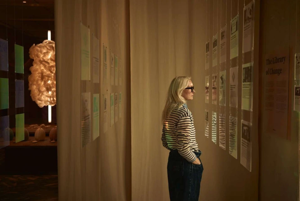 Woman in striped shirt examines informational panels at gallery exhibit with off-white walls and unique modern light fixture nearby.