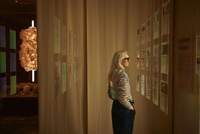 Woman in striped shirt examines informational panels at gallery exhibit with off-white walls and unique modern light fixture nearby.