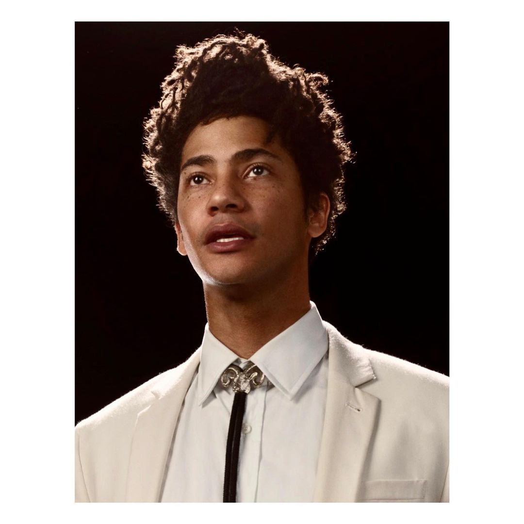 Headshot of a handsome adult male with curly hair, fair skin, wearing a white suit and bolo tie against a dark background.