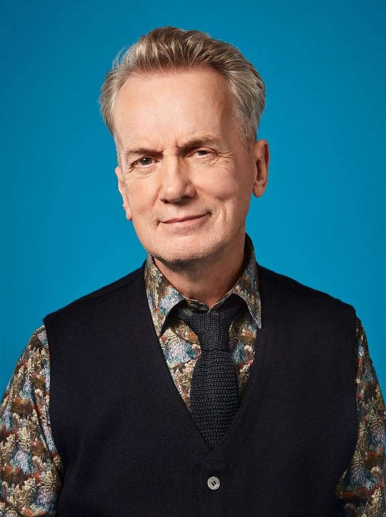 Headshot of an adult male in a patterned shirt and vest with a tie, posing against a blue background. The man has short grey hair.