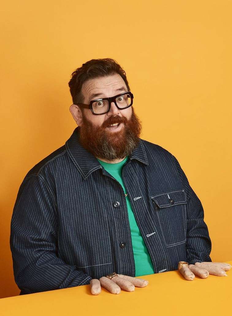 Man with red beard and glasses in front of orange backdrop, wearing denim jacket, smiling, with hands on table.