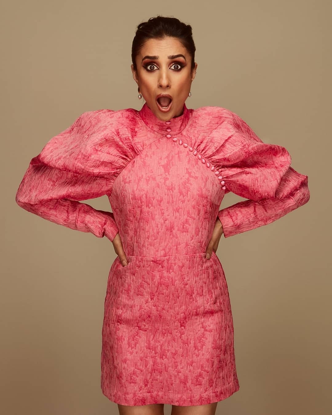 Portrait of a woman in a pink dress with exaggerated sleeves, hands on hips, against a neutral background. The woman has an open mouth.