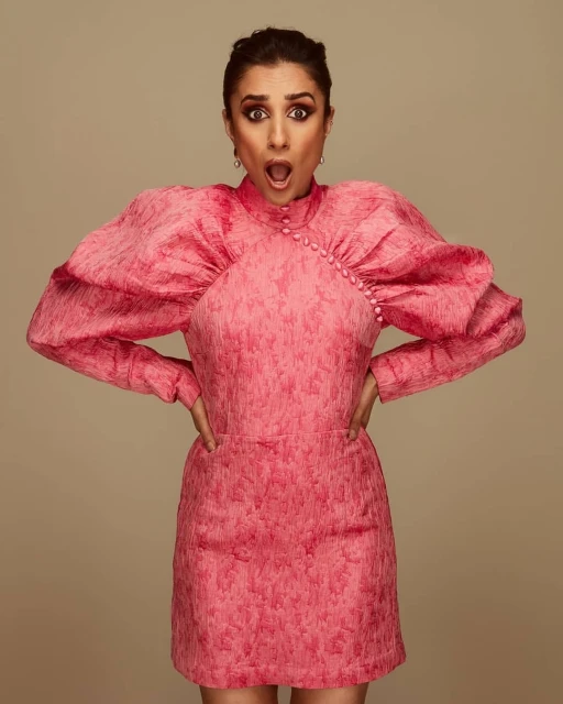 Portrait of a woman in a pink dress with exaggerated sleeves, hands on hips, against a neutral background. The woman has an open mouth.