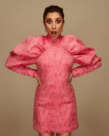 Portrait of a woman in a pink dress with exaggerated sleeves, hands on hips, against a neutral background. The woman has an open mouth.