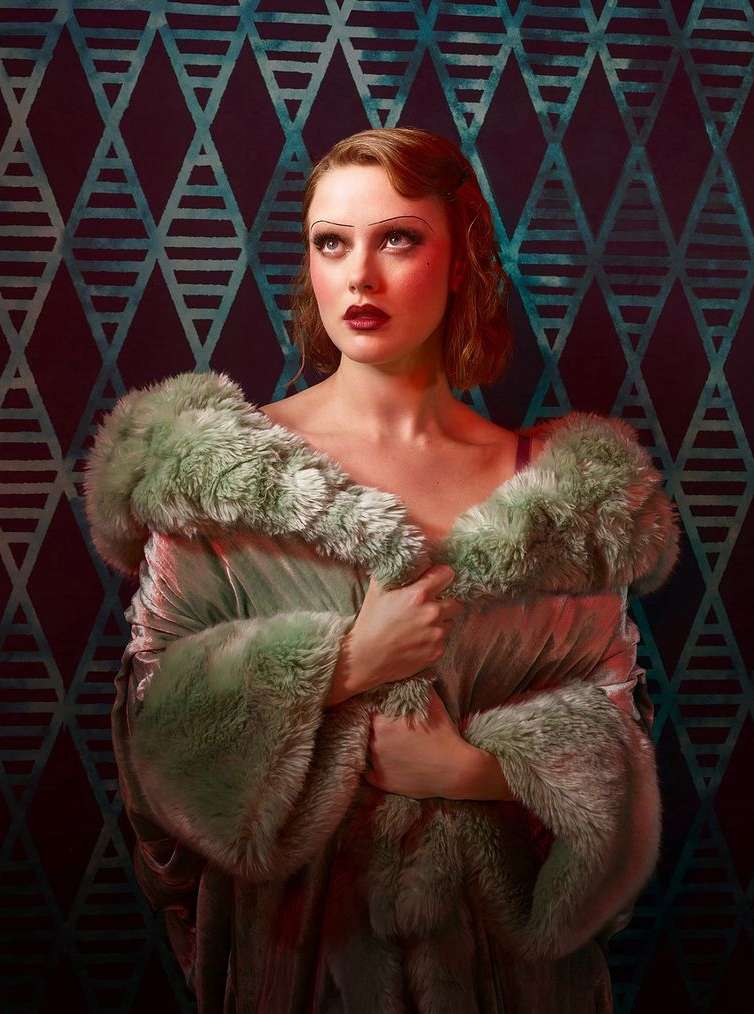 Portrait of a woman with dark red lipstick and vintage hair, wearing a light green fur coat against a blue and black patterned backdrop.