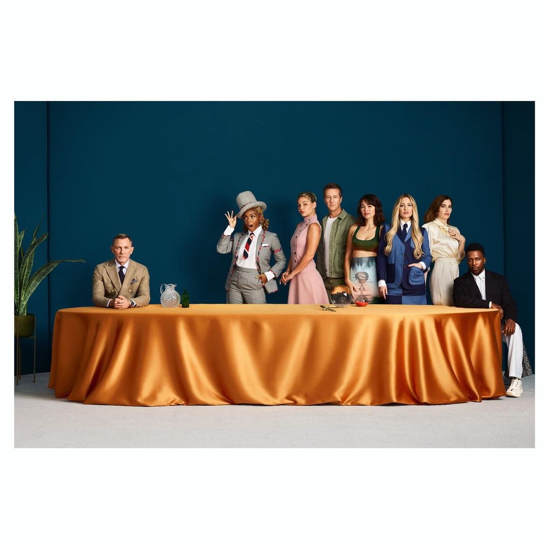 Group of seven people pose behind a long table with a gold tablecloth, against a dark teal backdrop in a studio setting.