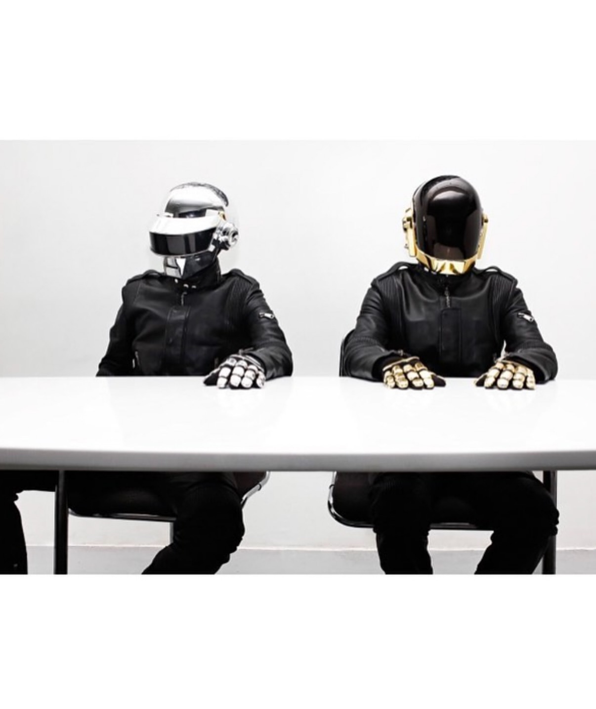 Two people wearing Daft Punk helmets and black leather jackets sit at a white table in a brightly lit, minimalist room. 
