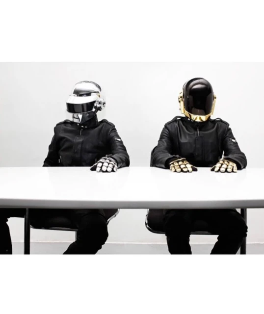 Two people wearing Daft Punk helmets and black leather jackets sit at a white table in a brightly lit, minimalist room. 