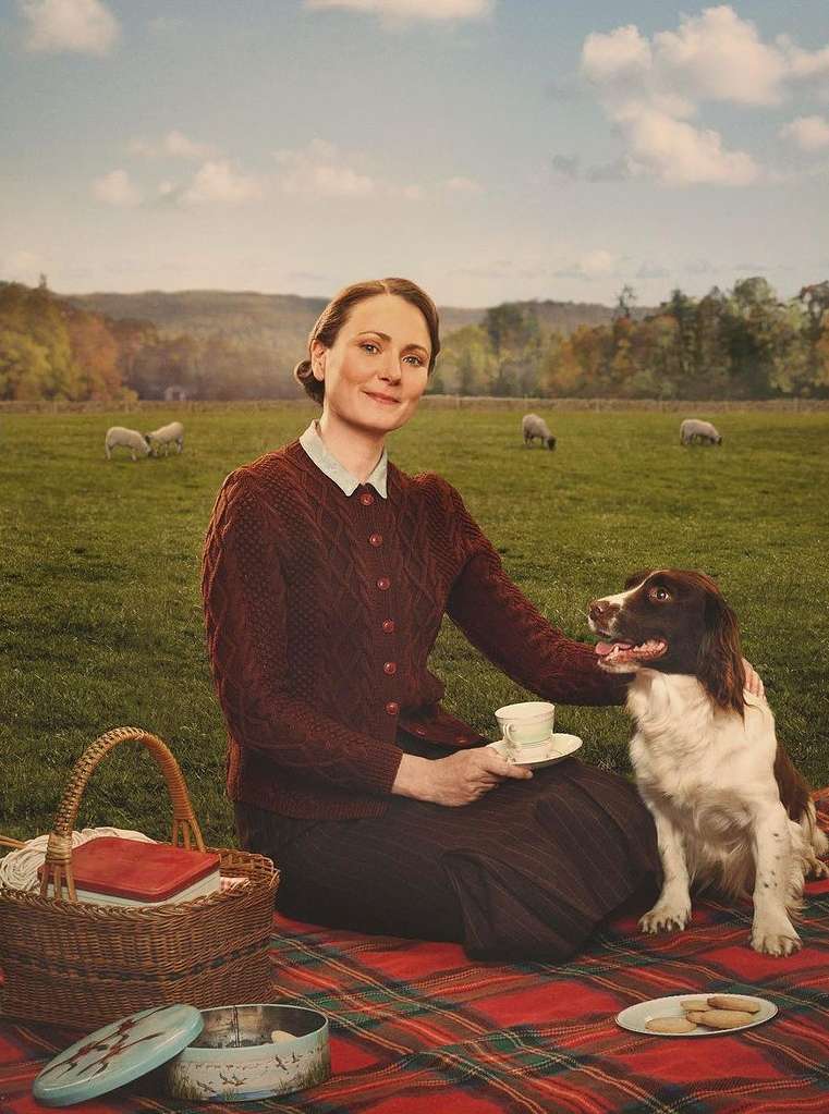 Woman and dog enjoy a picnic on a green field with sheep in the distance on a sunny day, creating a serene outdoor scene.